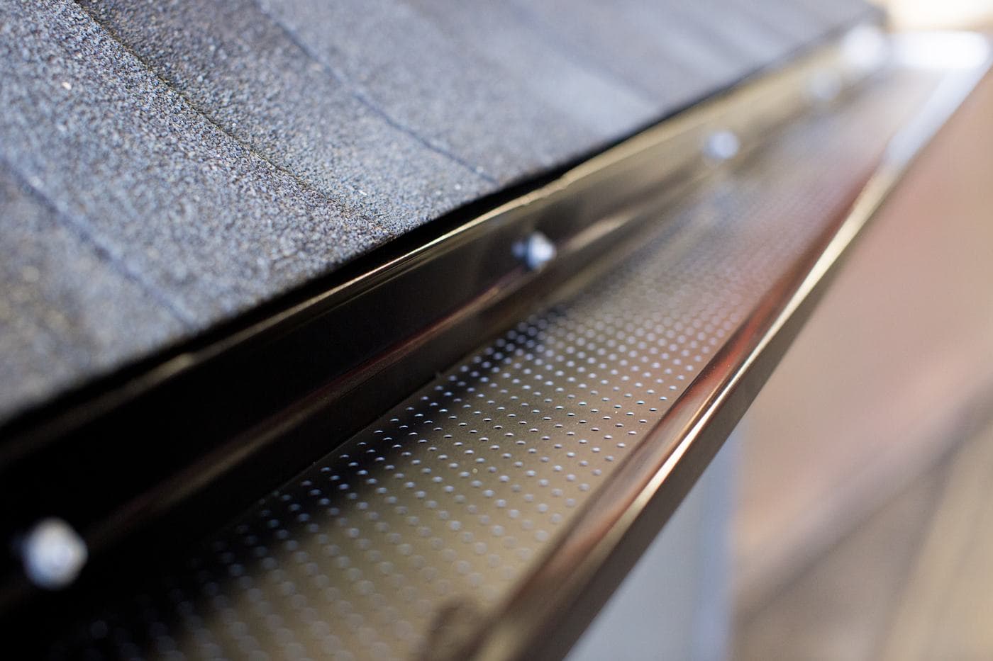 Close view of an installed gutter guard along a shingled roof edge, its drainage perforations open across the full run